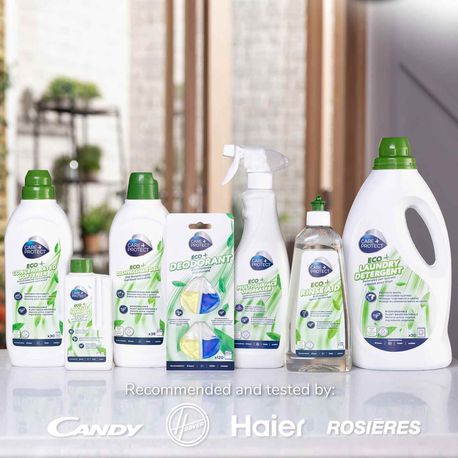 Cleaning products are neatly arranged on a reflective surface, including sprays and detergents with green and white packaging. Text reads: "Recommended and tested by: Candy, Haier, Rosières." Indoor setting with decorative plants.