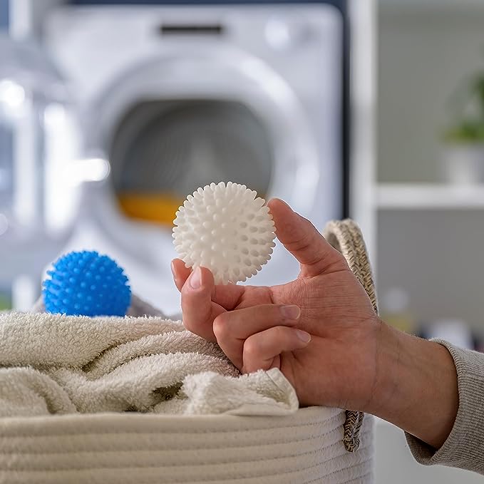 A hand holds a white spiky laundry ball over a basket filled with towels, with a blue ball and a washing machine visible in the background.