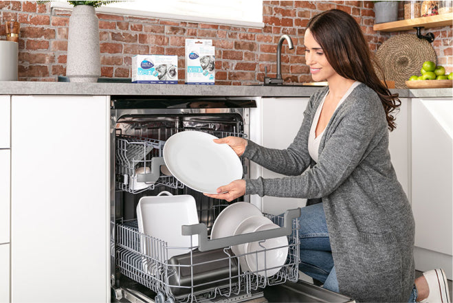 A woman places a white plate in an open dishwasher inside a modern kitchen with brick walls and a countertop displaying cleaning product boxes.