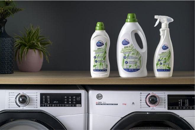 Laundry detergent bottles stand on a wooden surface above washing machines. Text: "Eco+ Laundry Detergent" and "Freshness." Two potted plants are placed on the left side.