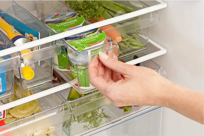 A hand opens a refrigerator drawer, revealing yogurts labeled "Schmand," carrots, and greens in a transparent organizer, amid other stored food items.
