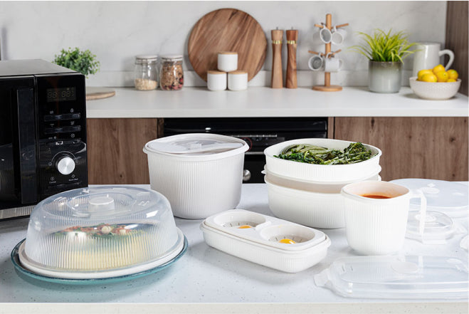 Plastic food containers sit on a kitchen counter, holding cooked vegetables and a beverage. Nearby, a microwave and various kitchen items like jars, spices, and a fruit bowl provide context.