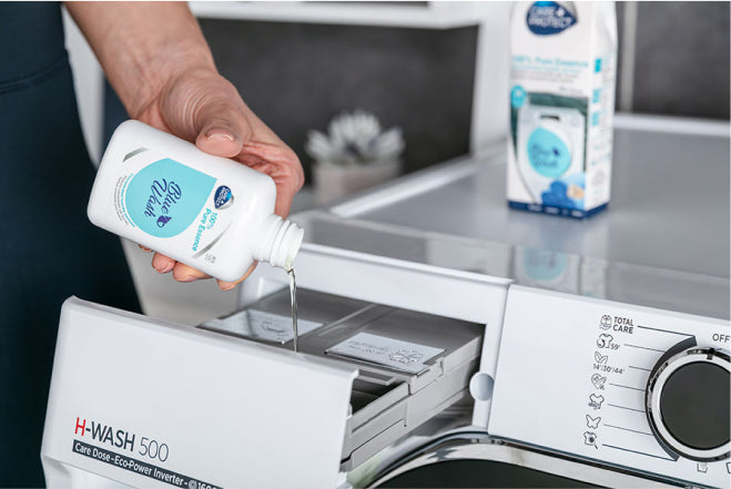 A hand pours liquid detergent from a bottle labeled "Eco Clean Care Dose" into a washing machine drawer. The machine, labeled "H-WASH 500," is in a modern laundry room.