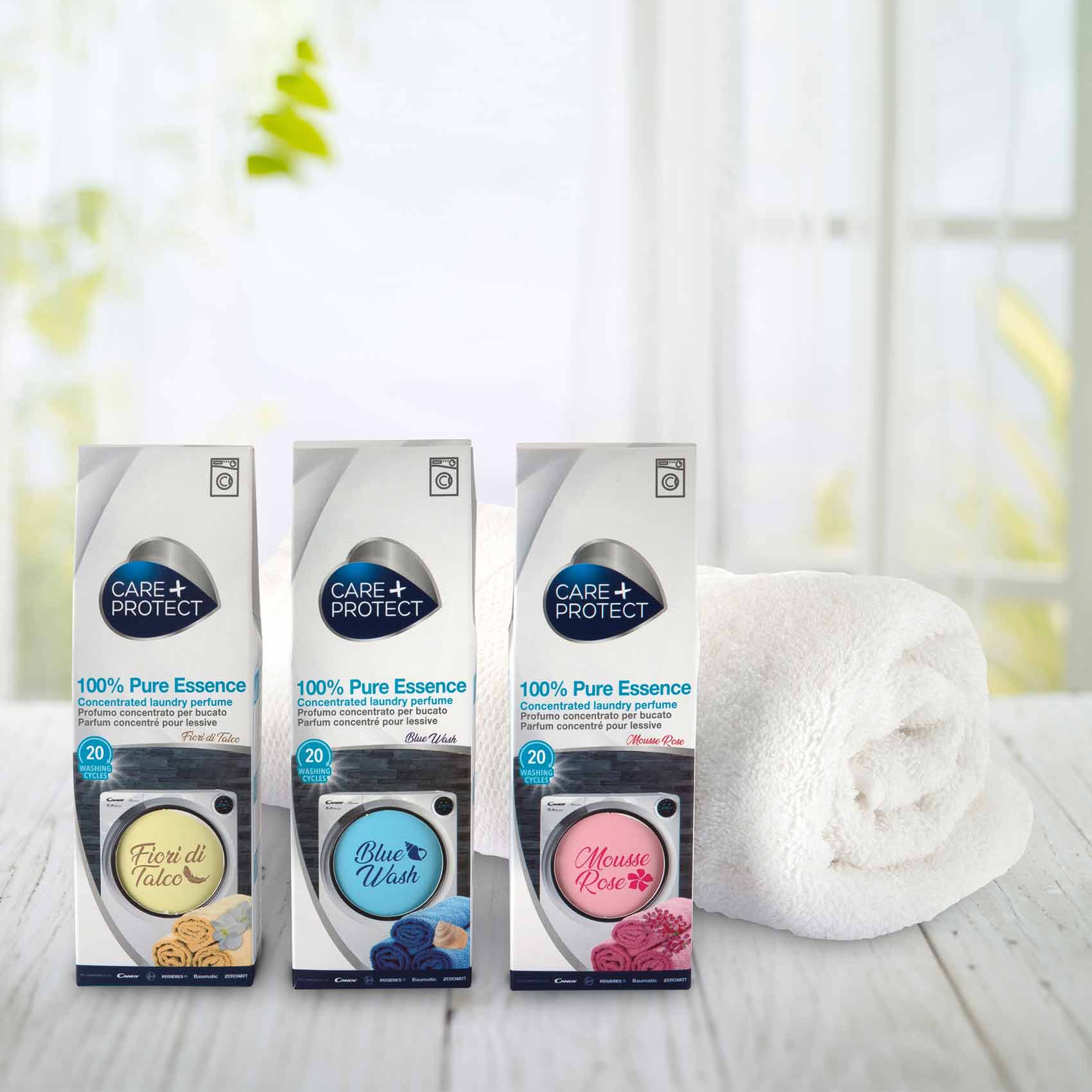 Three boxes of "Care + Protect 100% Pure Essence" laundry perfume stand on a wooden surface, each box featuring different scents: Fiori di Talco, Blue Wash, and Mousse Rose. A rolled white towel and a blurred window with greenery are in the background.