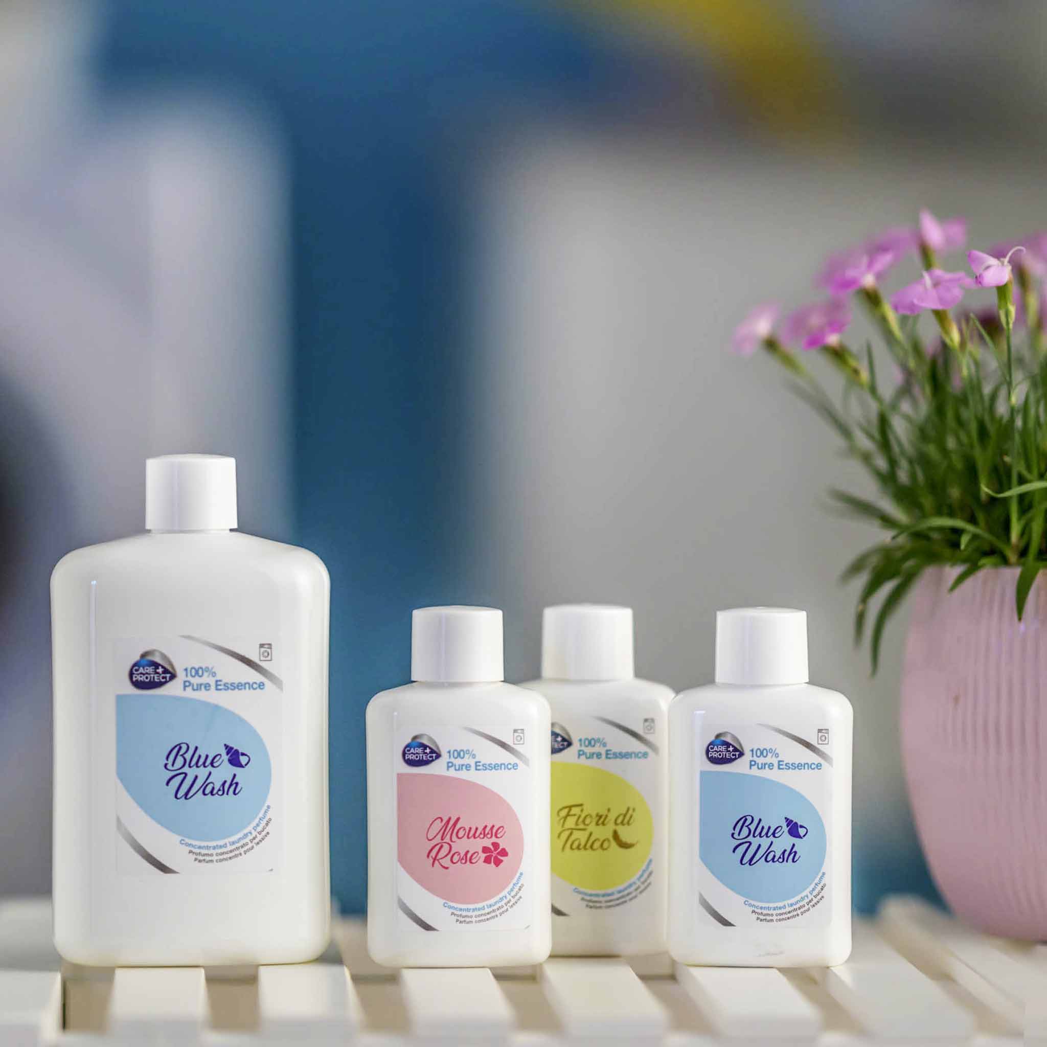 Bottles of "100% Pure Essence" products labeled Blue Wash, Mousse Rose, and Fiori di Talco sit on a white surface, with a pink pot of flowers nearby. Background is blurred blue and white.