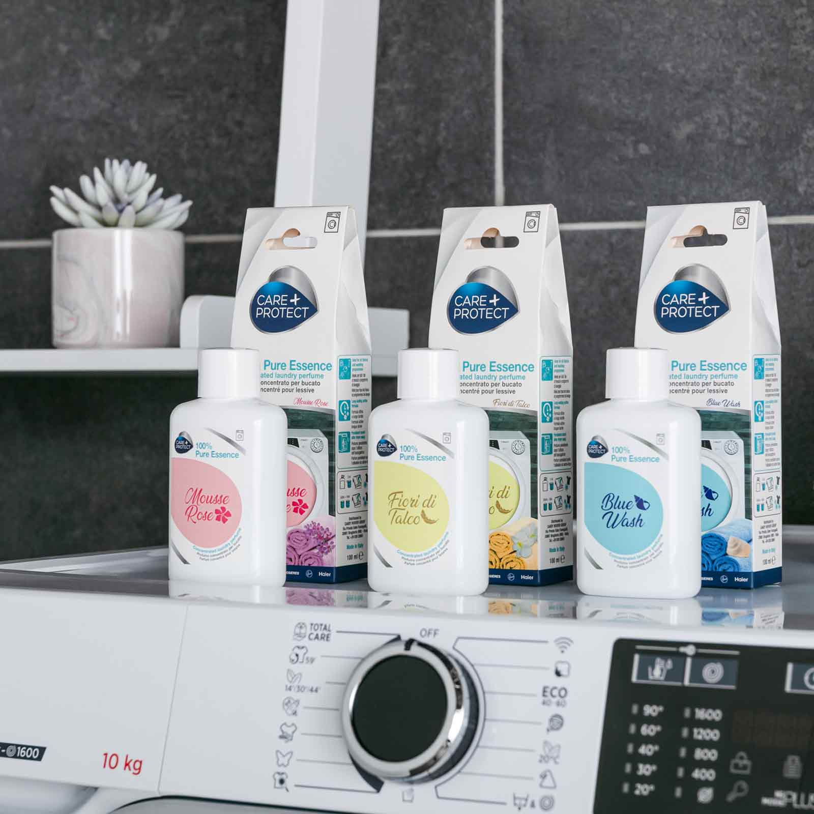 Laundry perfume bottles labeled "Mousse Rose," "Fiori di Talco," and "Blue Wash" are displayed on a washing machine. Behind them, "Care Protect Pure Essence" boxes are visible, with a potted plant on the left.