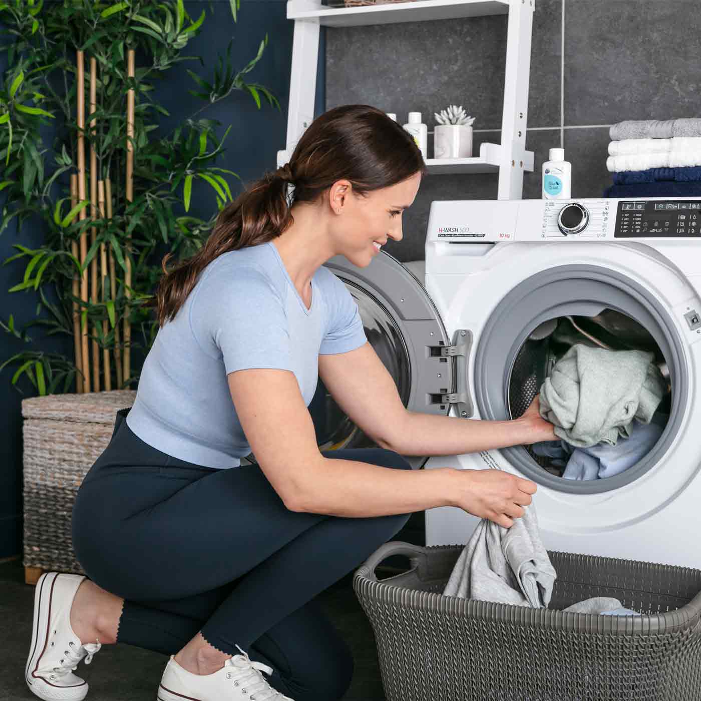 A woman crouches, placing clothes into a front-loading washing machine. The laundry room features a plant, wicker basket, and shelf with towels and detergent bottles.