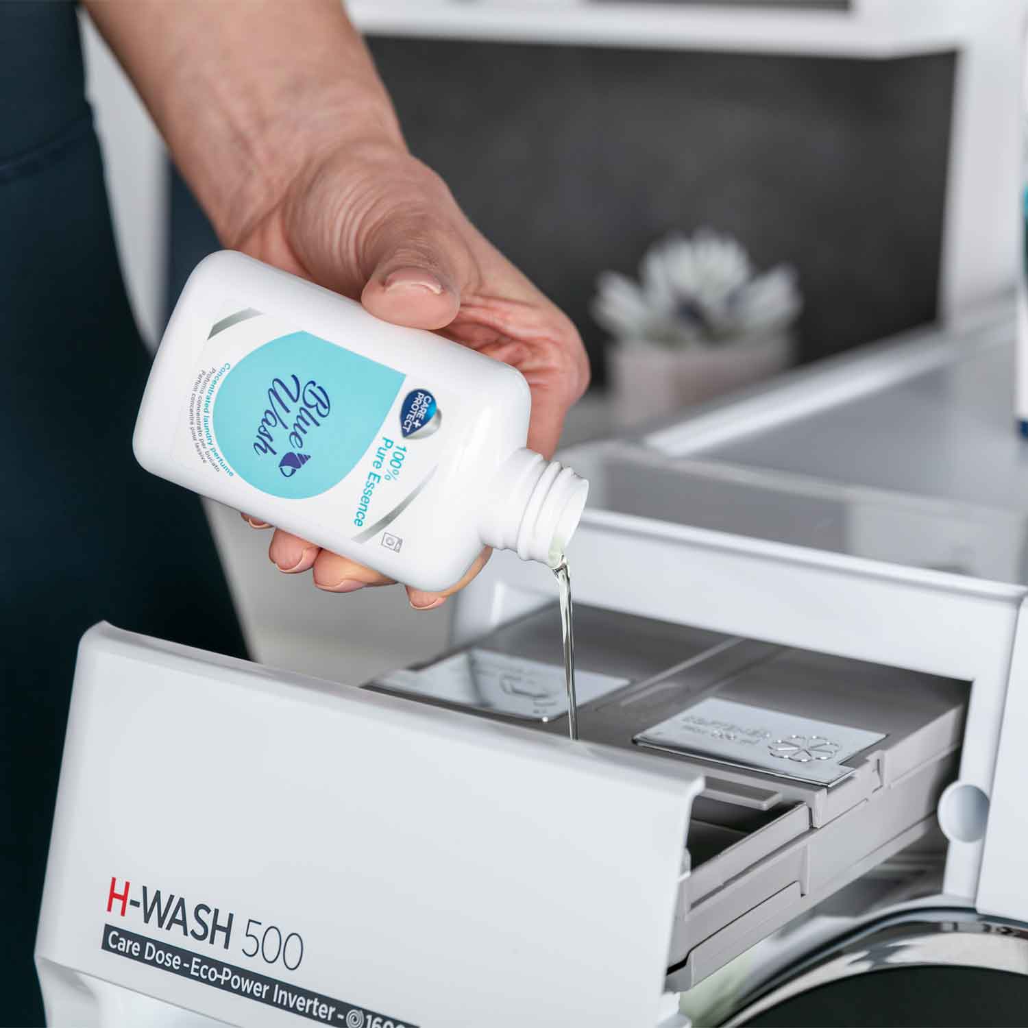 A hand pours liquid from a bottle labeled "Blue Wash, 100% Pure Essence" into an open washing machine drawer labeled "H-WASH 500, Care Dose - Eco-Power Inverter."