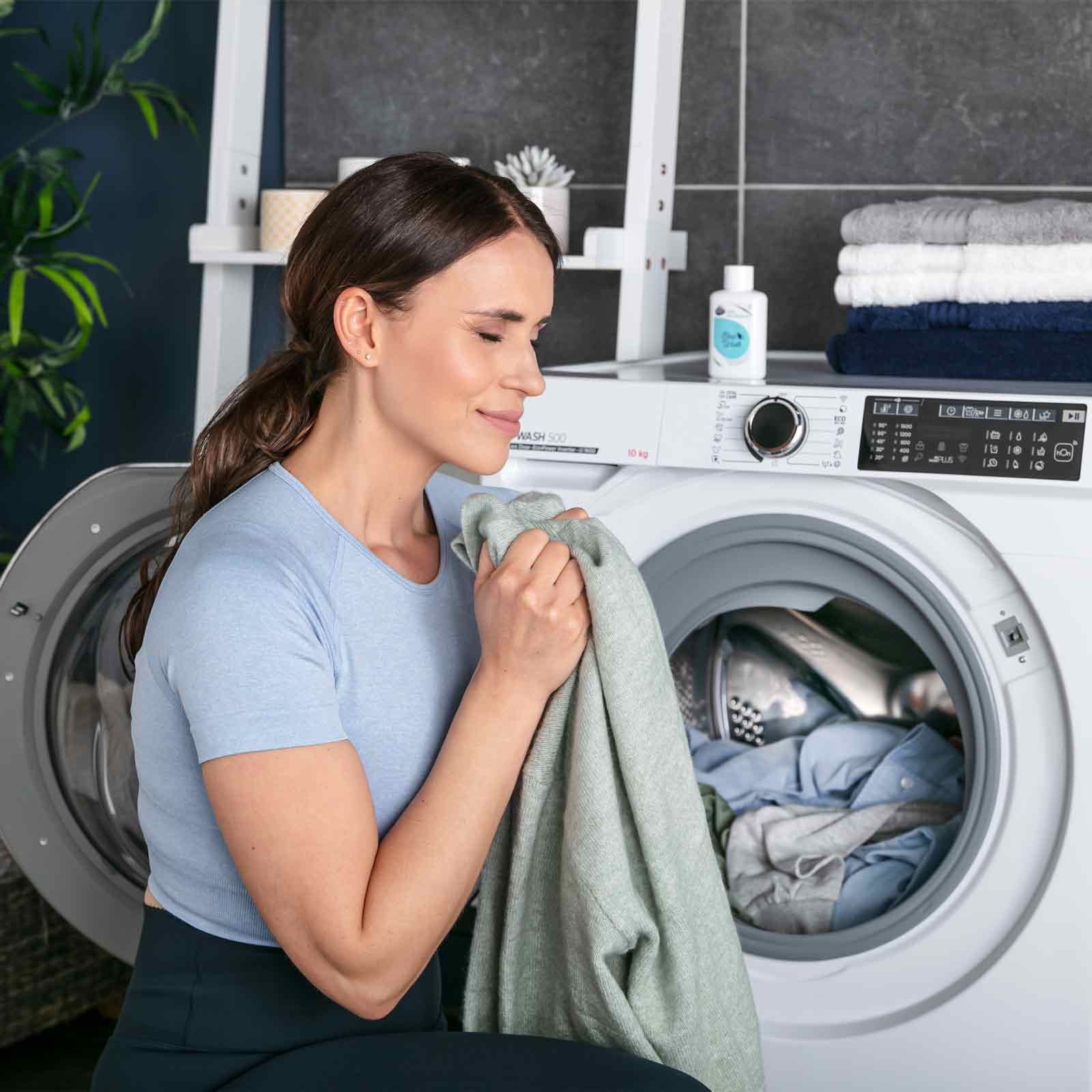 A woman in a light blue shirt smells a fresh garment near an open washing machine. Stacked towels and laundry detergent are placed on top, with plants in the background.